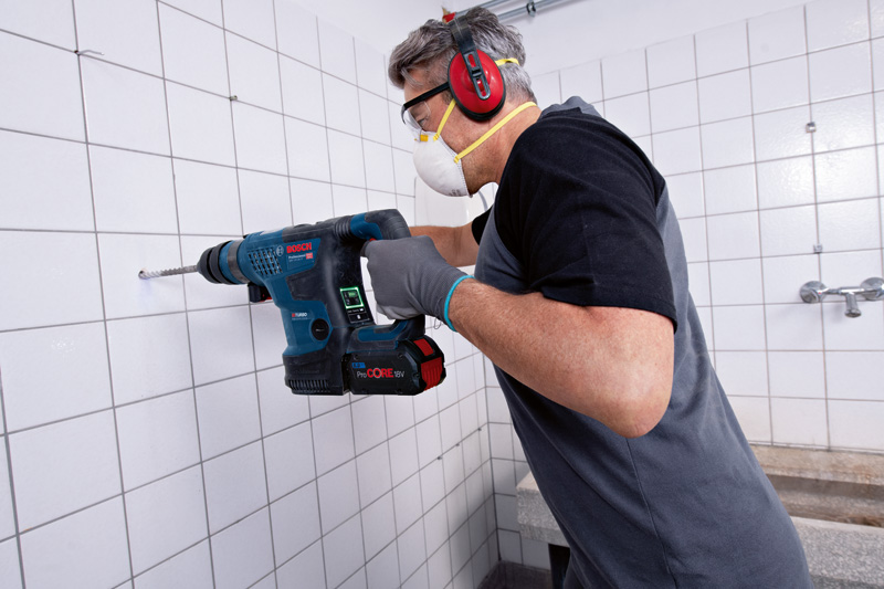 Eine Person mit Schutzausrüstung bohrt mit einem Akku-Bohrhammer in eine geflieste Wand. Anwendungsbild