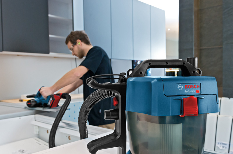 Eine Person mit Sicherheitsausrüstung verwendet einen Elektrohobel auf einem Holzbrett in der Nähe eines Staubsaugers. Anwendungsbild