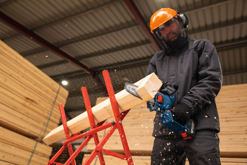 Eine Person mit Sicherheitsausrüstung schneidet im Innenbereich mit einer Akku-Hochentastersäge Holz. Anwendungsbild