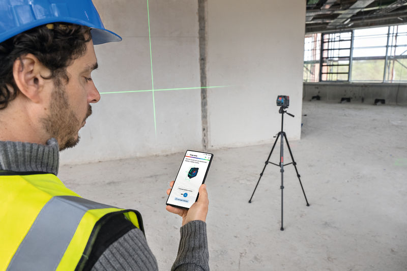 Eine Person mit Sicherheitsausrüstung steuert auf einer Baustelle ein Lasernivelliergerät mit einem Smartphone. Anwendungsbild
