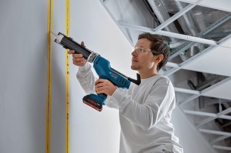 Eine Person mit Sicherheitsausrüstung trägt mit einer kabellosen Kartuschenpistole Dichtungsmittel auf eine Wand auf. Anwendungsbild