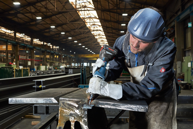 Eine Person mit Sicherheitsausrüstung bohrt in einer Fabrik in einen Metallträger. Anwendungsbild