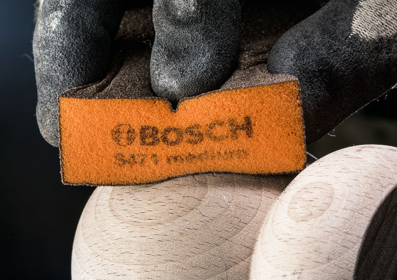 Behandschuhte Hand hält einen Schleifblock gegen eine glatte Holzoberfläche und trägt Sicherheitsausrüstung. Anwendungsbild