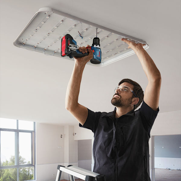 Eine Person mit Sicherheitsausrüstung installiert mit einer Bohrmaschine eine Deckenleuchte. Anwendungsbild