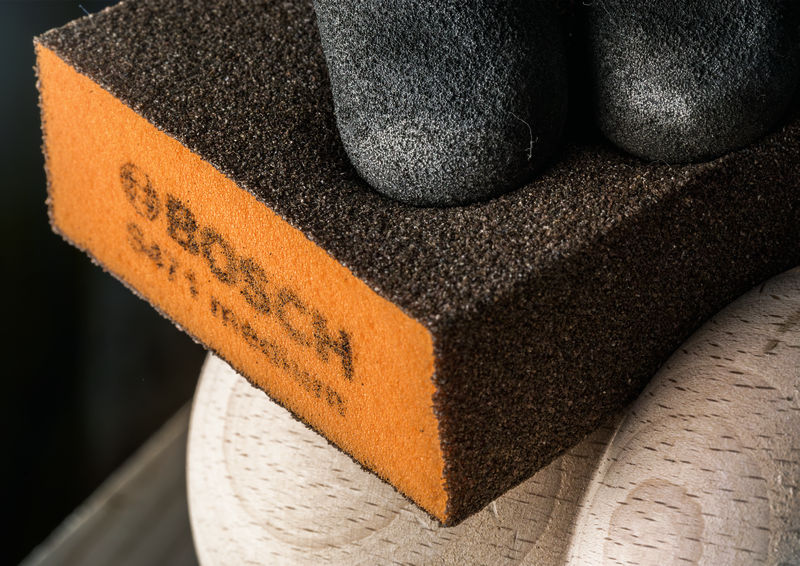 Eine Hand in einem schwarzen Handschuh hält einen Schleifschwamm gegen eine Holzoberfläche. Anwendungsbild